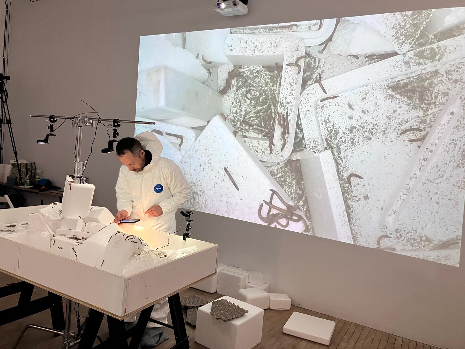 August Black in white protective suit standing in front of a large white table with lights on it and a project of superworms behind him.